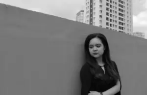 Young woman in black attire leaning against a wall, with urban buildings in the background. Black and white photography.