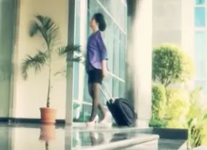 Woman with a suitcase walking towards a modern building entrance, surrounded by greenery and a potted palm tree.