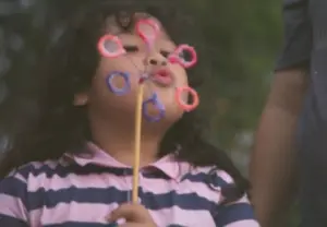 Child blowing bubbles with a bubble wand, wearing a striped shirt, enjoying a sunny day outdoors.