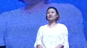 Woman in a white top stands confidently on stage, with a blue background highlighting her presence at a public event.