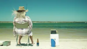 Person fishing on a beach, sitting in a chair with a cooler and beer, enjoying a sunny day by the water.