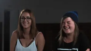 Two young women laughing together, showcasing a cheerful moment in a dimly lit environment.