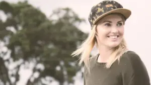 Smiling woman in a camo cap stands outdoors, with trees softly blurred in the background capturing a cheerful atmosphere.