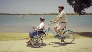 An elderly woman enjoys a ride in a bike trailer while a man pedals along a scenic waterfront path on a sunny day.