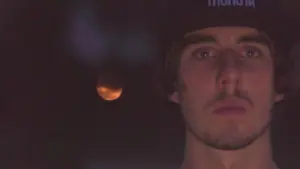 Young man with intense gaze, wearing a black cap, with a blurred orange moon in the background at night.