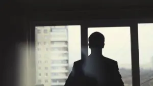 Silhouette of a man in a suit standing by a window, overlooking a city skyline on a cloudy day.