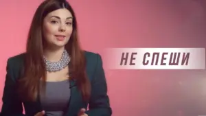 Woman in a green blazer with a statement necklace, speaking against a pink backdrop, conveying a message of patience.