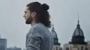 Man with curly hair in a stylish sweater stands against a city skyline with historic buildings in the background.