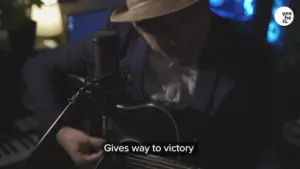 Musician in a hat performing live with a guitar, creating a poignant atmosphere in a dimly lit setting.