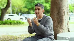 Man in gray sweater gestures while seated outdoors, surrounded by trees and blurred cars in the background.