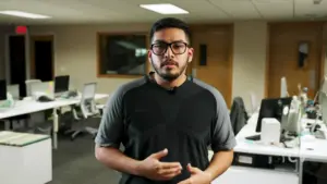 Man with glasses standing in a modern office environment, surrounded by desks and computers.