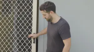 Man opening a door with a security mesh screen, dressed casually and looking down, in a residential setting.