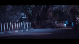 Dimly lit street at night, featuring a parked car and shadows cast by trees, creating an eerie, atmospheric scene.