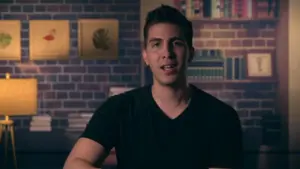 Man in a black shirt sitting in front of a bookshelf, with framed art on the wall, against a cozy brick backdrop.