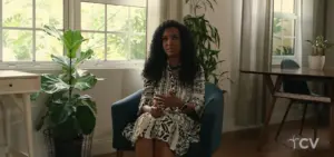 Woman sitting in a cozy room with plants, wearing a patterned dress, engaged in conversation, emphasizing a serene atmosphere.