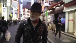 Stylish man in a black leather jacket and fedora walking through a bustling urban street at night.