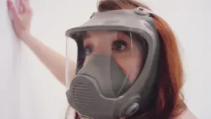 Woman wearing a protective respirator mask, looking up against a white wall, emphasizing safety and health awareness.