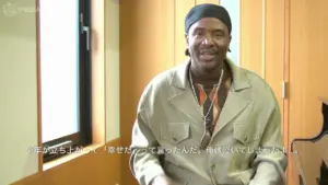 Man sitting indoors, wearing a stylish outfit and headscarf, with a warm and inviting background.