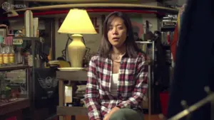 Woman sitting in a cozy room with a lamp, wearing a plaid shirt, surrounded by books and art.