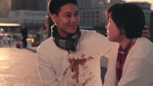 Couple sitting together outdoors, smiling, with a city skyline backdrop and a playful chocolate stain on the man's shirt.