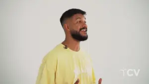 Man with a beard wearing a yellow shirt speaking enthusiastically against a plain white background.