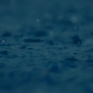 Close-up of raindrops splashing on a dark, reflective surface, creating ripples in the water.