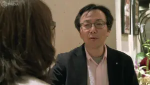 A man in glasses and a blazer converses with a woman, both engaged in a casual discussion.