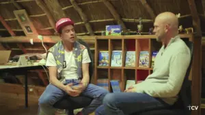 Two men engaged in conversation inside a cozy, rustic room with bookshelves and cultural decor.