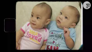 Two adorable babies sitting side by side, wearing colorful shirts with cheerful messages, smiling and playful expressions.