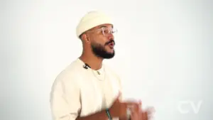 Man wearing glasses and a beanie gestures enthusiastically while speaking against a plain white background.