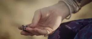 A close-up of a hand delicately holding a small, ornate ring against a blurred natural background.