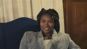 Smiling woman with curly hair sitting in an armchair, dressed in a stylish coat, with a cozy indoor background.