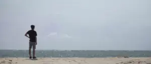 Person standing on a sandy beach, gazing at the calm ocean under a cloudy sky.