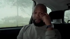 Man sitting in a car during rain, looking contemplative while tapping his finger to his temple.