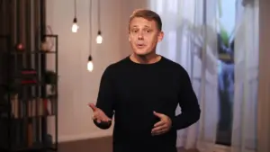 Man in a black sweater gestures while speaking in a modern room with hanging lights and a window backdrop.