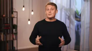 Man in a black sweater gestures while speaking in a well-lit room with hanging light bulbs and a bright background.