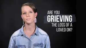 Woman in a denim shirt asking if viewers are grieving the loss of a loved one, against a dark background.