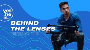 A filmmaker crouches with a camera on a gimbal against a blue background, promoting the "Behind the Lenses" series.