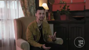 Man sitting in an armchair, speaking expressively in a warmly lit room with bookshelves and indoor plants.