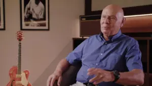 Elderly man in a blue shirt seated in a cozy room with guitar and framed photos on the wall, conveying a reflective mood.