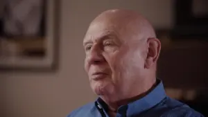 Elderly man with a bald head and blue shirt, thoughtfully looking to the side in a softly lit indoor setting.