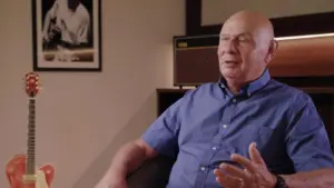 An older man in a blue shirt sits in a cozy room with a guitar and a framed photo in the background.