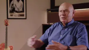 Older man in a blue shirt discusses topics, guitar in background, framed photo on wall, warm lighting, indoor setting.