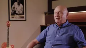 A man seated in a blue shirt, with a guitar and framed photo of a baseball player in a cozy room setting.