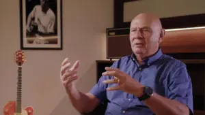 Older man in a blue shirt gestures while discussing a topic, with a guitar and framed photo in the background.