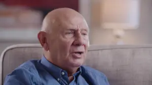 Elderly man with a bald head, wearing a blue shirt, seated on a couch, engaged in conversation against a soft background.