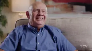 Smiling older man in blue shirt sitting comfortably on a couch, enjoying a relaxed moment indoors.