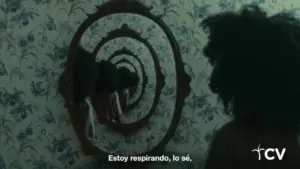 A person with curly hair gazes into a spiraling mirror reflection on floral wallpaper, evoking a sense of introspection.