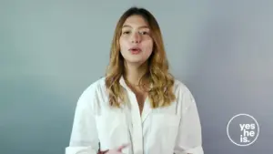 Woman with brown wavy hair wearing a white shirt, speaking confidently against a gray background for a motivational campaign.