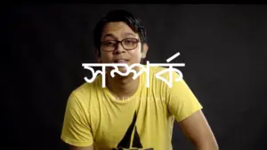 Young man in a yellow t-shirt with glasses, smiling against a dark background featuring Bengali text.